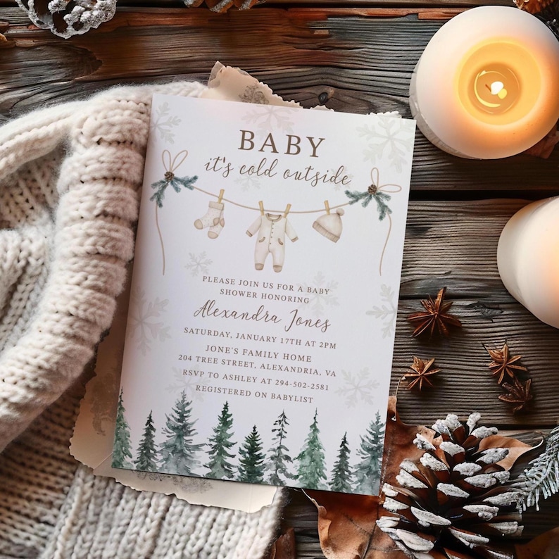 May include: A baby shower invitation with the text "BABY it's cold outside." The invitation features illustrations of baby clothes hanging on a clothesline, snowflakes, and a forest scene. The invitation is surrounded by candles and pinecones.