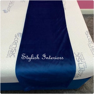 May include: A dark blue velvet table runner with the text "Stylish Interiors" embroidered in white on the front. The runner is laying on a white mattress with a pattern of small dots.