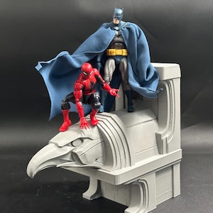 Könnte beinhalten: Eine rot-blaue Spider-Man-Actionfigur hockt auf einer grauen, steinartigen Adler-Wasserspeier-Statue, während eine dunkelblau gekleidete Batman-Actionfigur mit langem blauem Umhang und gelbem Gürtel dahinter auf einem grauen Sockel steht.