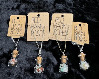 May include: Four silver necklaces with glass bottles filled with colorful stones. Each necklace has a tag with the text "Red Barn Roses" and a different word below it: "Protection", "Manifest", "Heat the Hole", and "Joy".