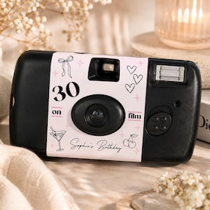 Op de afbeelding: Een zwarte wegwerpcamera met een op maat gemaakte lichtroze papieren wikkel met de tekst 30 on film en Sophie's Birthday. De wikkel bevat zwarte lijntekeningen van een strik, hartjes, een martiniglas, kersen en kleine sterretjes als accenten.