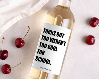 Op de afbeelding: Een fles witte wijn met een etiket met de tekst "Turns Out You Weren't Too Cool For School." Er liggen verschillende kersen rond de fles. De achtergrond is licht en neutraal.