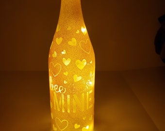 May include: A glass bottle with a gold glitter finish and white hearts and the text "bee mine" on the front. The bottle is lit from inside, creating a warm glow.