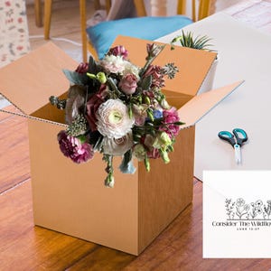 May include: A bouquet of fresh flowers in a brown cardboard box. The flowers are a mix of pink, white, and purple blooms. A pair of scissors and a card with the text "Consider The Wildflowers" are also visible.