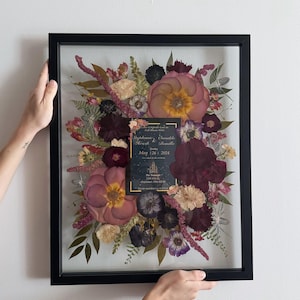 May include: A framed pressed flower arrangement with a wedding invitation card in the center. The invitation card is gold and black with the text "Stephanie & Brendan" and "May 12th, 2024".