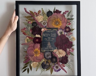 May include: A framed pressed flower arrangement with a wedding invitation card in the center. The invitation card is gold and black with the text "Stephanie & Brendan" and "May 12th, 2024".