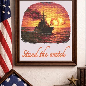 May include: Cross-stitch artwork in a wooden frame depicting a warship at sunset, with the text "Stand the watch". The image features a vibrant orange and yellow sky, and a folded American flag.