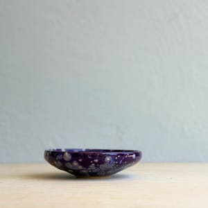 May include: A small, round ceramic bowl featuring a deep purple glaze with a speckled, crystalline pattern in shades of white, light blue, and grey. The bowl has a smooth, glossy finish and a low-profile, shallow design with curved edges.