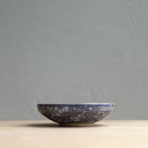 May include: A small, round ceramic bowl with a speckled blue and gold glaze. The bowl has a low profile and sits on a light-colored surface. The background is a solid, muted gray.