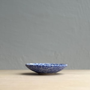 以下が含まれることがあります： 青と白の斑点模様の小さな丸い陶器のボウル。ボウルは深さが浅く、縁がわずかに広がっています。ボウルは、少量の食べ物を提供したり、装飾品として使用したりする可能性があります。