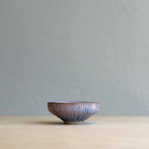 May include: A small ceramic bowl with a tapered base and a wide, open rim. The exterior features a mottled glaze with a blend of soft pink, lavender, and deep blue streaks that run vertically down the sides toward the bottom of the vessel.
