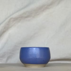 Könnte beinhalten: Eine kleine, handgefertigte Keramikschale mit mattem periwinkelfarbenem Blau außen und einem natürlichen, unglasierten beigefarbenen Boden. Die Schale hat eine abgerundete Form mit einem leicht facettierten Design. Die gesprenkelte Textur sorgt für visuelles Interesse.