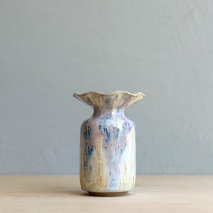 May include: A ceramic vase with a ruffled, flared rim and a cylindrical body. The surface features a mottled glaze with blended tones of pale pink, soft blue, and creamy yellow, creating a vertical drip effect across the exterior of the vessel.