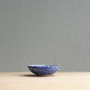 May include: A small, round ceramic bowl with a speckled blue and white pattern. The bowl has a slightly flared rim and a light beige base. The bowl is set against a neutral background, highlighting its simple design.
