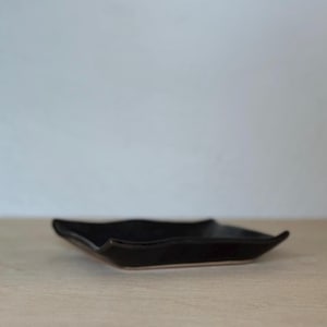 May include: A small, black ceramic dish with a unique, wavy shape. The dish has a matte finish and sits on a light-colored surface. The design is simple and modern, suitable for serving small portions or as a decorative accent.