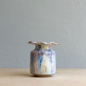 May include: A small ceramic vase with a ruffled, wavy rim. The body features a reactive glaze with vertical streaks of blue, lavender, and cream colors over a light base. The base of the vase is unglazed, showing the natural tan clay texture.