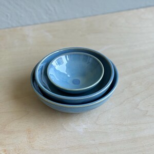 May include: Three nesting bowls in shades of blue, arranged in a stack. The bowls have a glossy finish and a slightly textured appearance. The rims of the bowls have a lighter, almost cream-colored edge. The bowls are on a light wood surface.