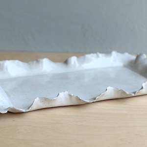 May include: A rectangular ceramic tray with a glossy white finish and subtle brown speckles. The tray features an uneven, wavy, and organic edge that mimics the appearance of folded or rippled material, creating a distinct textured border.