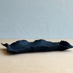 May include: A rectangular ceramic tray with a matte black finish and an irregular, wavy, hand-formed edge. The surface of the tray features a subtle speckled texture, and the overall form is elongated with a low profile and a flat base.