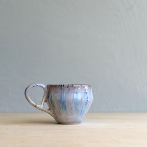 May include: A ceramic mug with a wide, tapered body and a single handle. The exterior features a reactive glaze with vertical streaks of light blue, soft pink, and cream tones, transitioning to a darker, brownish rim at the top edge.
