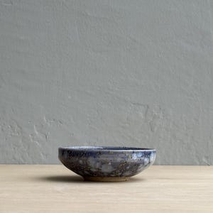 May include: A small, handmade ceramic bowl with a speckled blue and gray glaze. The bowl has a rounded shape and sits on a light-colored surface. The background is a textured gray wall. This bowl is suitable for serving small portions.