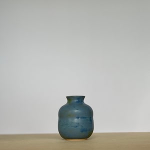 Puede incluir: Un pequeño jarrón de cerámica azul con cuerpo redondeado y abertura ligeramente acampanada. El jarrón tiene una superficie moteada y texturizada.