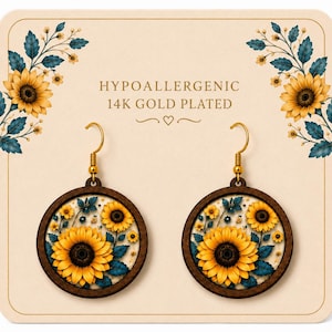 May include: A pair of round wooden earrings featuring a sunflower design. The earrings have gold-colored hooks and are set against a cream-colored background. The text "HYPOALLERGENIC 14K GOLD PLATED" is printed on the background.