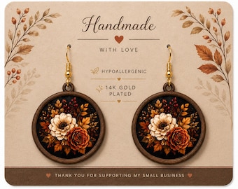 May include: A pair of round wooden earrings featuring a floral print with cream and orange flowers on a black background. The earrings have gold-toned hooks and are displayed on a tan card with the text Handmade with love, hypoallergenic, 14k gold plated.