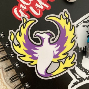 Puede incluir: Una pegatina de un fénix morado y amarillo que se eleva de las llamas. El fénix es blanco con un contorno gris. La pegatina está en una libreta negra.
