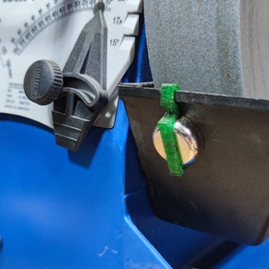 Può includere: Una mola da affilare blu e nera con una guida angolare verde. La mola da affilare ha un quadrante bianco con segni per i gradi e una manopola nera. La guida angolare è fissata alla mola da affilare con una vite argentata.