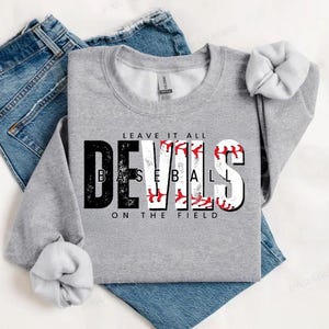 May include: A gray sweatshirt with the text "Leave it all" above the word "Devils" in black and white with a baseball graphic. The text "Baseball" is written in white letters with red stitching on the baseball graphic. The text "On the field" is written below the graphic.