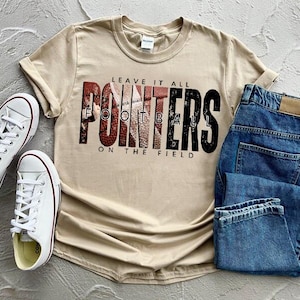 May include: A casual outfit featuring a tan t-shirt with the text "LEAVE IT ALL POINTERS FOOTBALL ON THE FIELD", blue jeans, and white sneakers with red accents. The shirt has a crew neck and rolled sleeves.