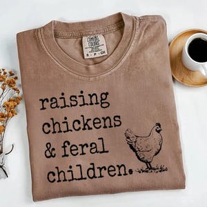May include: A brown t-shirt with the text "raising chickens & feral children." printed in black. A black and white illustration of a chicken is to the right of the text.