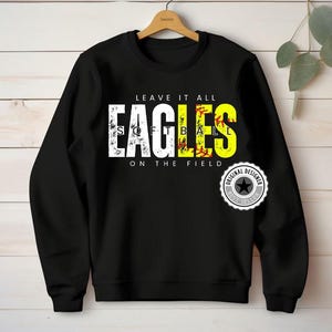 Puede incluir: Sudadera negra con el texto "LEAVE IT ALL EAGLES SOFTBALL ON THE FIELD" en blanco y amarillo. El diseño incluye un gráfico de softbol y un logotipo de "ORIGINAL DESIGNER BEST SELLER".