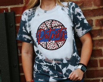 May include: White t-shirt with a blue and white tie-dye pattern. The shirt features a red and black basketball with the word "Patriots" written in red and "Basketball" written in black.
