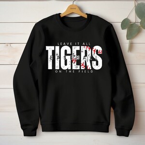 May include: Black sweatshirt with white text that reads "Leave it all Tigers Baseball on the field". The word "Tigers" is in a large font and has a red baseball graphic inside the letters.