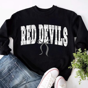 Puede incluir: Sudadera negra con la inscripción "RED DEVILS" en blanco con estampado de camuflaje. Un lazo gris está debajo del texto. La sudadera se combina con vaqueros azules y zapatillas blancas con detalles rojos. Una pequeña planta verde en el fondo.