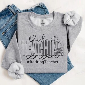 K&ouml;nnte beinhalten: Ein graues Sweatshirt mit dem Text "the last teaching season #RetiringTeacher" in Schwarz. Neben dem Text ist ein Stapel B&uuml;cher abgebildet.