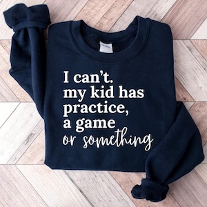 May include: Navy blue sweatshirt with white text that reads: "I can't. my kid has practice, a game or something." The sweatshirt has a crew neck and long sleeves with ribbed cuffs. The garment is folded.
