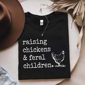 May include: Black t-shirt with white text that reads "raising chickens & feral children." A white silhouette of a chicken is to the right of the text.