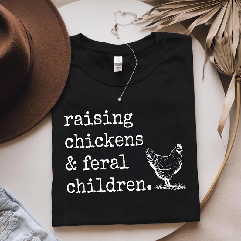 May include: Black t-shirt with white text that reads "raising chickens & feral children." A white silhouette of a chicken is to the right of the text.