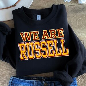 May include: Black sweatshirt with the text "WE ARE RUSSELL" in gold and maroon lettering. The sweatshirt is folded, with a portion of blue jeans visible at the bottom. A white hat is in the background.