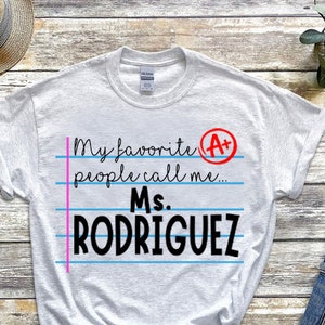 May include: A white t-shirt with a pink and blue lined design that says "My favorite people call me... Ms. RODRIGUEZ" with an A+ circled in red.