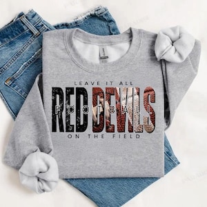 May include: A grey sweatshirt featuring the text "LEAVE IT ALL RED DEVILS FOOTBALL ON THE FIELD" in black and red. The sweatshirt is folded on top of a pair of blue jeans.