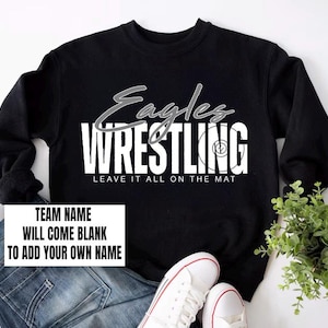 May include: Black sweatshirt with the text "Eagles Wrestling" in white and grey. The phrase "LEAVE IT ALL ON THE MAT" is below the word "Wrestling". A white rectangle at the bottom says "TEAM NAME WILL COME BLANK TO ADD YOUR OWN NAME".