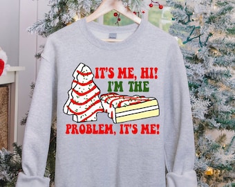 May include: A light gray sweatshirt with the text "IT'S ME, HI! I'M THE PROBLEM, IT'S ME!" in red and green. The graphic includes a Christmas tree and a cake slice with red and green accents.