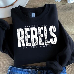May include: Black sweatshirt with the text "LEAVE IT ALL REBELS ON THE COURT" in distressed white lettering. The word "REBELS" features a basketball design. The sweatshirt is folded on a wooden surface.