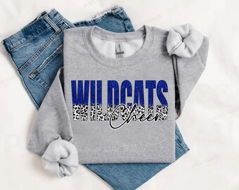 Peut inclure: Un sweat-shirt gris avec l'inscription "Wildcats Cheer" imprimée sur le devant. Le texte est en paillettes bleues avec un imprimé léopard noir et blanc en dessous.