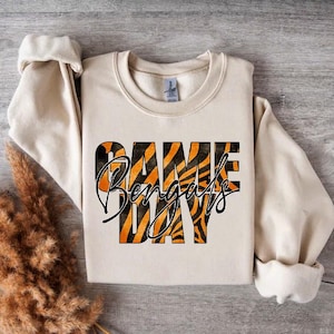 Puede incluir: Una sudadera de color crema con un dise&ntilde;o de rayas de tigre que dice "Game Day Bengals" en letras negras y naranjas.