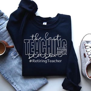 K&ouml;nnte beinhalten: Ein marineblauer Sweatshirt mit dem Text "The Last Teaching Season #RetiringTeacher" in Wei&szlig;. Der Text ist von einem Stapel B&uuml;cher umgeben.